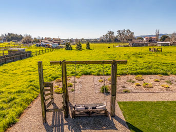 Wooden hanging bench swing on a gravel patio overlooking a sunlit green pasture, fenced farmland and distant farm buildings under a clear blue sky.
