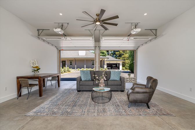 Finished two-car garage converted into a cozy living area in a suburban home with open garage doors showing the driveway; features a gray sofa with blue pillows, leather accent chair, round wire coffee table on a patterned area rug, wooden desk with chair, large modern ceiling fan, polished concrete floor, and potted orchids.