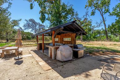 Sunny backyard rustic outdoor kitchen pavilion with covered bar and corrugated metal siding, a covered grill and stainless gas grill, string lights, picnic table with closed umbrella, and oak trees under a clear blue sky