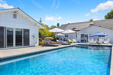 Sunny suburban backyard with a sparkling rectangular pool, concrete patio, white ranch-style house with solar panels, lounge chairs, cushions and striped umbrellas