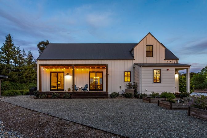 Modern farmhouse exterior at dusk — cozy covered porch with warm string lights, gravel driveway, Adirondack chairs on wooden steps, raised garden beds and surrounding trees.