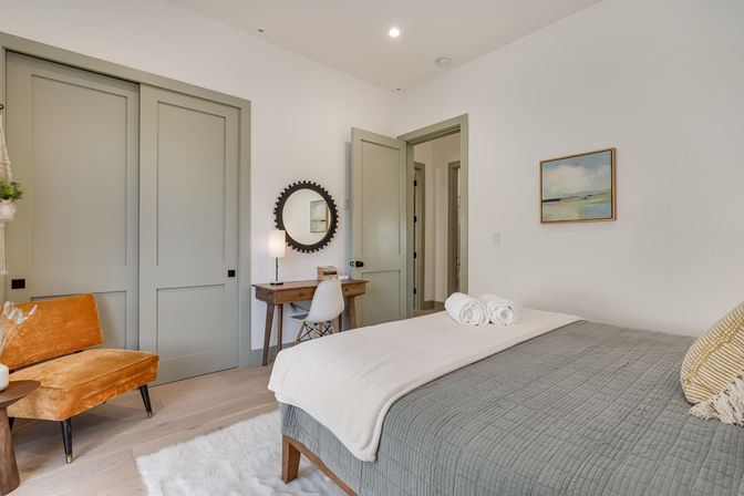 Bright modern bedroom with gray quilted bed and rolled towels, wooden desk with round mirror, orange velvet accent chair, pale green sliding closet doors and light wood floors