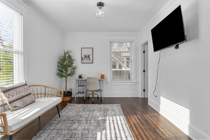 Bright sunlit home office with white walls and hardwood floors, wall-mounted TV, rattan bench, small desk with rolling chair, potted plant and large window blinds