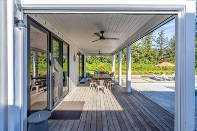 Inviting sunny covered patio with wood decking, sliding glass doors, ceiling fans, and a colorful outdoor dining table set; poolside lounge chairs and umbrella on a concrete terrace with green hedges and trees beyond.