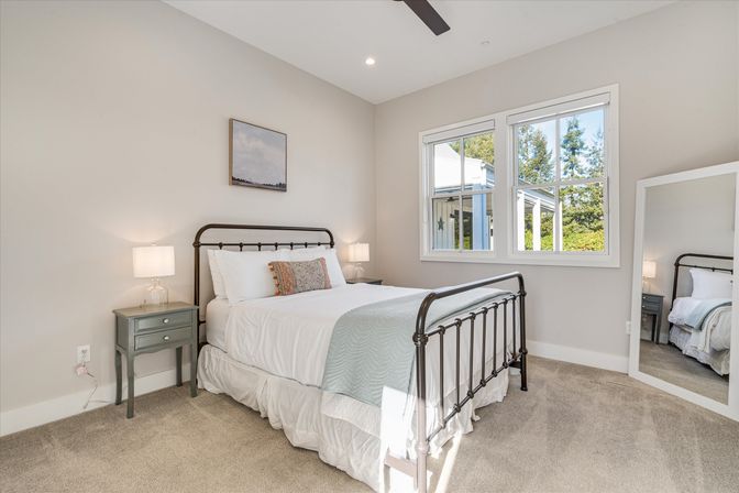 Sunlit suburban bedroom with black metal bed frame, white linens and pale blue throw, gray nightstands with lamps and a window showing green trees