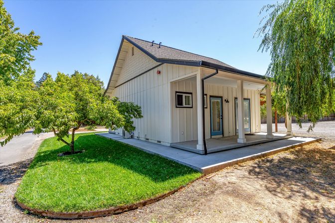 Bright white modern farmhouse exterior with covered front porch, blue door, small manicured lawn and leafy trees under a clear blue sky.