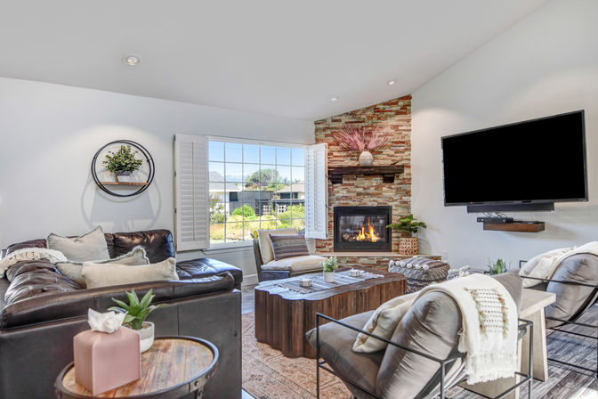 Cozy sunlit living room with stone fireplace, leather sectional, wood coffee table, large shuttered window and wall-mounted TV