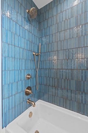 Coastal-inspired bathroom with glossy blue vertical tiles surrounding a white bathtub, brushed-bronze showerhead, handheld wand, tub spout and matching controls.