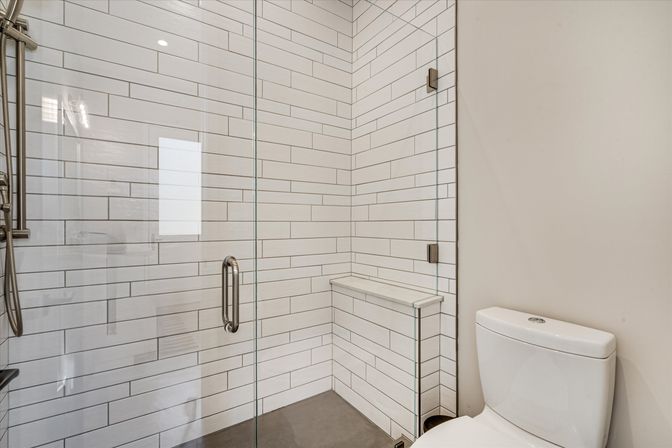 Sleek modern bathroom with a frameless glass walk-in shower clad in white horizontal subway tile, a built-in corner bench, brushed metal fixtures, and an adjacent white toilet.