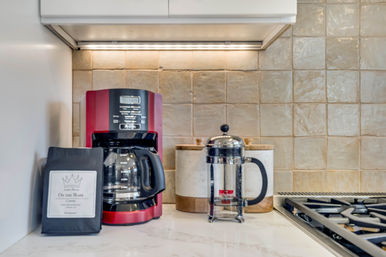 Kitchen countertop coffee station with a red drip coffee maker and glass carafe, French press, marble canisters and beige tiled backsplash beside a gas cooktop