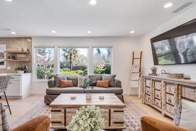 Bright modern farmhouse living room with large windows overlooking a lush garden, gray sofa with rust accent pillows, rustic wood coffee table and media console, wall-mounted TV and open kitchen bar area.