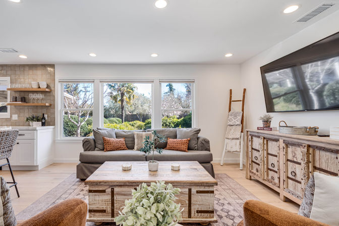 Bright modern farmhouse living room with large windows overlooking a lush garden, gray sofa with rust accent pillows, rustic wood coffee table and media console, wall-mounted TV and open kitchen bar area.
