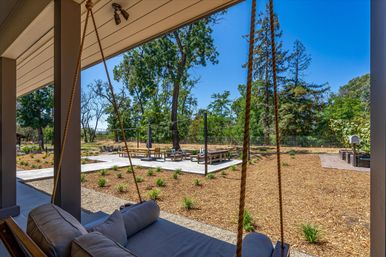 View from a shaded porch with a rope-hung cushioned swing overlooking a sunny landscaped backyard and outdoor living space — concrete patio with seating, wood-chip groundcover, young plantings and tall trees under a clear blue sky.