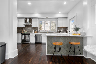 Bright modern open-concept kitchen with white cabinets, dark subway-tile backsplash, stainless steel appliances, marble-topped island with two wooden bar stools, pendant light and dark hardwood floors.