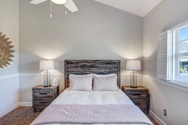 Bright cozy bedroom with rustic reclaimed-wood headboard, white linens and pale pink quilt, matching wood nightstands with textured glass lamps, ceiling fan and shuttered window letting in sunlight.