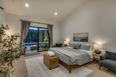 Contemporary mountain-view bedroom with king bed in gray linens, sliding glass doors opening to a patio fire pit, cozy chairs, wooden trunk and plush rug.