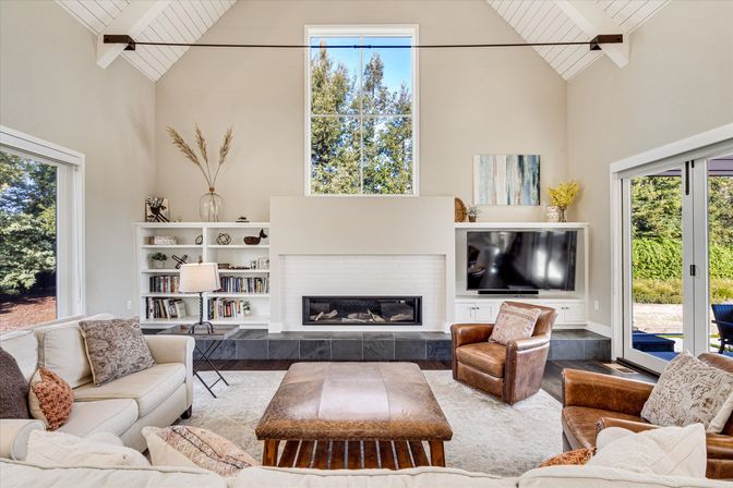 Airy modern farmhouse living room with vaulted white ceiling and tall window above a white-brick fireplace, built-in shelves and TV, beige sectional, leather armchairs and ottoman, and sliding glass doors opening to a tree-lined backyard.