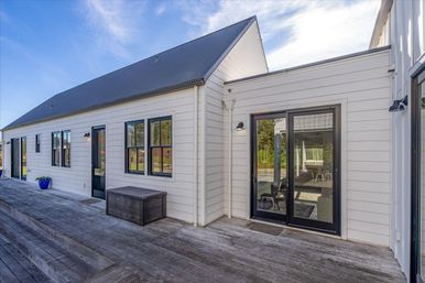 Sunlit white clapboard house exterior with black metal roof, black-framed windows and sliding glass doors opening onto a weathered wooden deck with a storage bench and blue planter under a clear blue sky.
