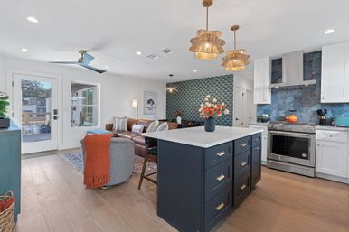 Open-concept coastal-modern kitchen and living area with a navy island and white quartz countertop, blue scalloped tile accent wall, rattan pendant lights, stainless steel range, leather sectional sofa, and light hardwood floors.