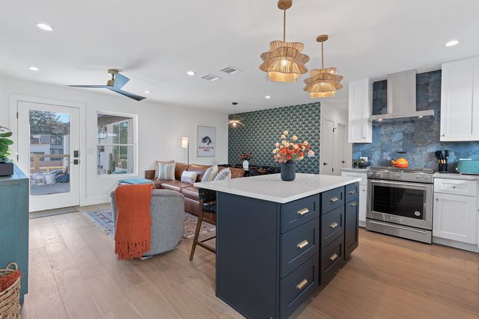 Open-concept coastal-modern kitchen and living area with a navy island and white quartz countertop, blue scalloped tile accent wall, rattan pendant lights, stainless steel range, leather sectional sofa, and light hardwood floors.