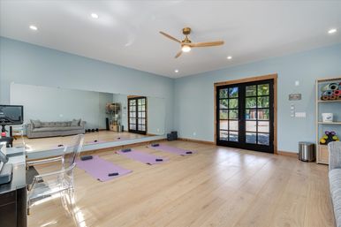 Bright, serene home yoga studio with light oak floors, mirrored wall, ceiling fan, three purple yoga mats and blocks, clear acrylic chair, gray sofa, shelving with rolled mats, and black French doors opening to a sunny yard.