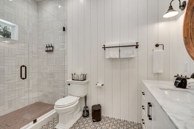 Bright modern farmhouse bathroom with white shiplap walls, patterned black-and-white floor tile, glass walk-in shower with light gray tiles, pedestal-style toilet, marble vanity, round wood mirror and bronze fixtures.