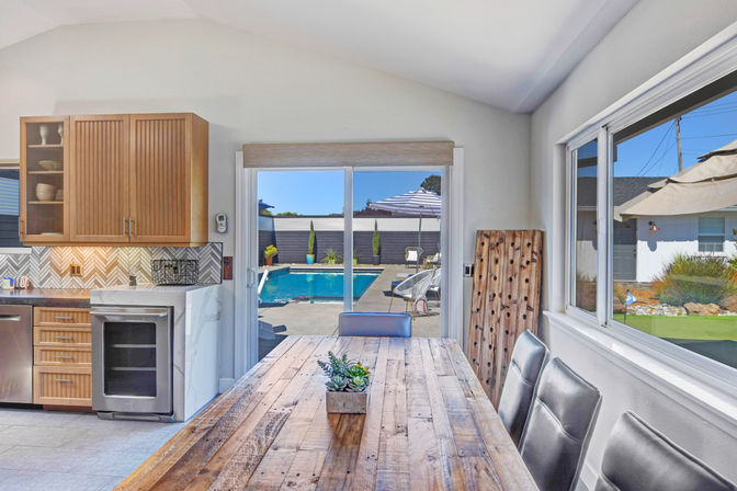 Bright modern kitchen-dining area with a rustic wooden table and leather chairs, sliding glass door opening to a sunny backyard patio and swimming pool with umbrella
