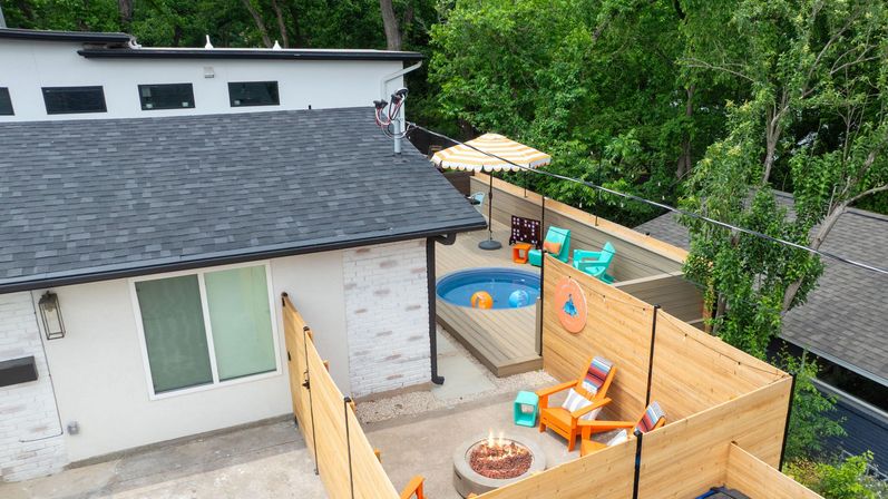 Cozy residential backyard deck with wooden privacy fence, small round plunge pool, striped umbrella, bright Adirondack chairs and a glowing fire pit.