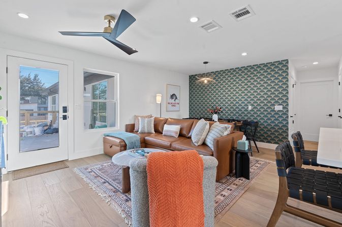 Bright modern open-plan living room with a caramel leather sectional, teal scallop accent wall, round coffee table, woven rug, ceiling fan, and hardwood floors