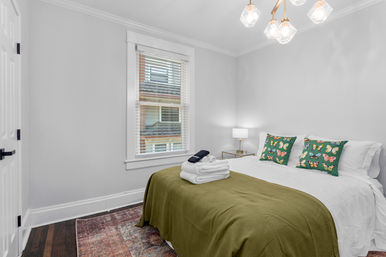 Bright modern apartment bedroom with white walls, queen bed dressed in white linens and olive-green throw, butterfly print pillows, folded towels on bed, bedside lamp, window blinds and hardwood floors.
