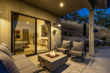 Covered hillside backyard patio at dusk with modern cushioned sofa and swivel chairs around a square gas fire pit, concrete floor and sliding glass doors revealing a cozy, warmly lit living room.