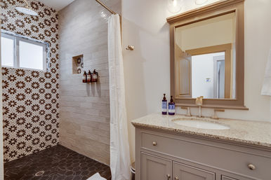 Spa-ready residential master bathroom with floral-pattern shower tiles, rain showerhead, dark hexagon floor, granite vanity and framed mirror.