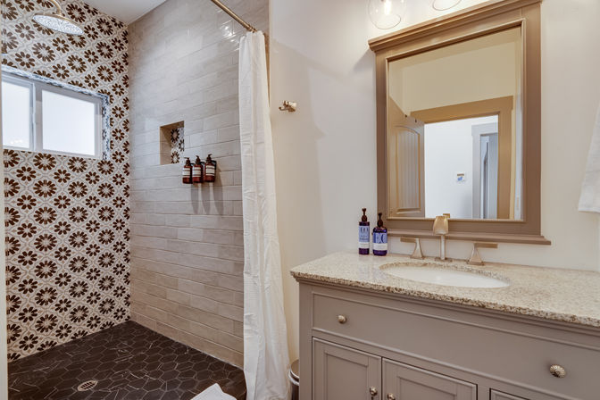 Spa-ready residential master bathroom with floral-pattern shower tiles, rain showerhead, dark hexagon floor, granite vanity and framed mirror.