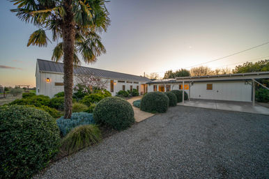 White ranch-style home with metal roof and carport, palm tree and neatly trimmed round shrubs lining a gravel driveway and walkway at sunset.