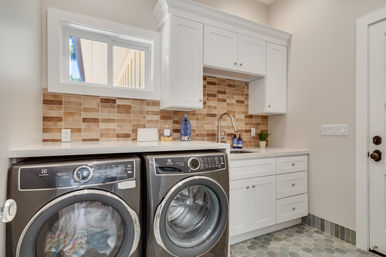 Bright modern laundry room with matching front-loading washer and dryer, white shaker cabinets and countertop, beige tile backsplash, stainless faucet and utility sink, hexagon tile floor, and small window above the work surface.