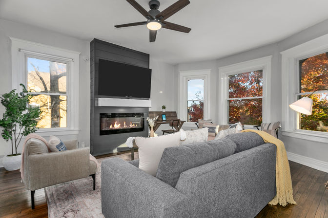 Cozy contemporary living room with gray sofa and armchair, gas fireplace topped by a wall-mounted TV, bay windows showing autumn foliage, hardwood floors, ceiling fan and a yellow throw