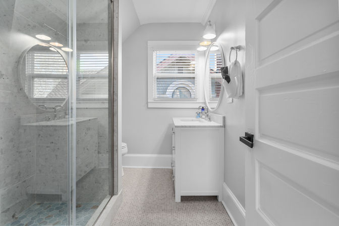 Bright modern residential bathroom with glass-enclosed marble shower and blue hex floor tiles, white vanity with round mirror and wall sconce, window with blinds showing rooftops, towel on a ring and an open paneled door.