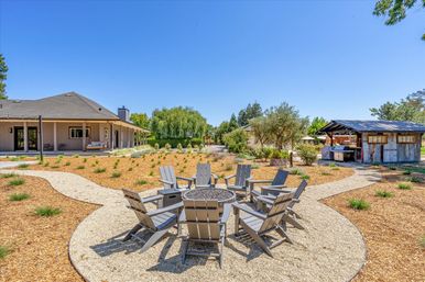 Sunlit residential backyard with a circular fire pit surrounded by gray Adirondack chairs on a gravel patio, winding paths through drought-tolerant landscaping, farmhouse porch to the left and rustic outdoor pavilion to the right under a clear blue sky.