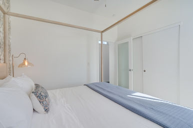 Sunlit minimalist bedroom with light-wood four-poster bed, crisp white linens, patterned accent pillow, blue quilted throw, glass pendant bedside lamp, and white sliding closet doors.