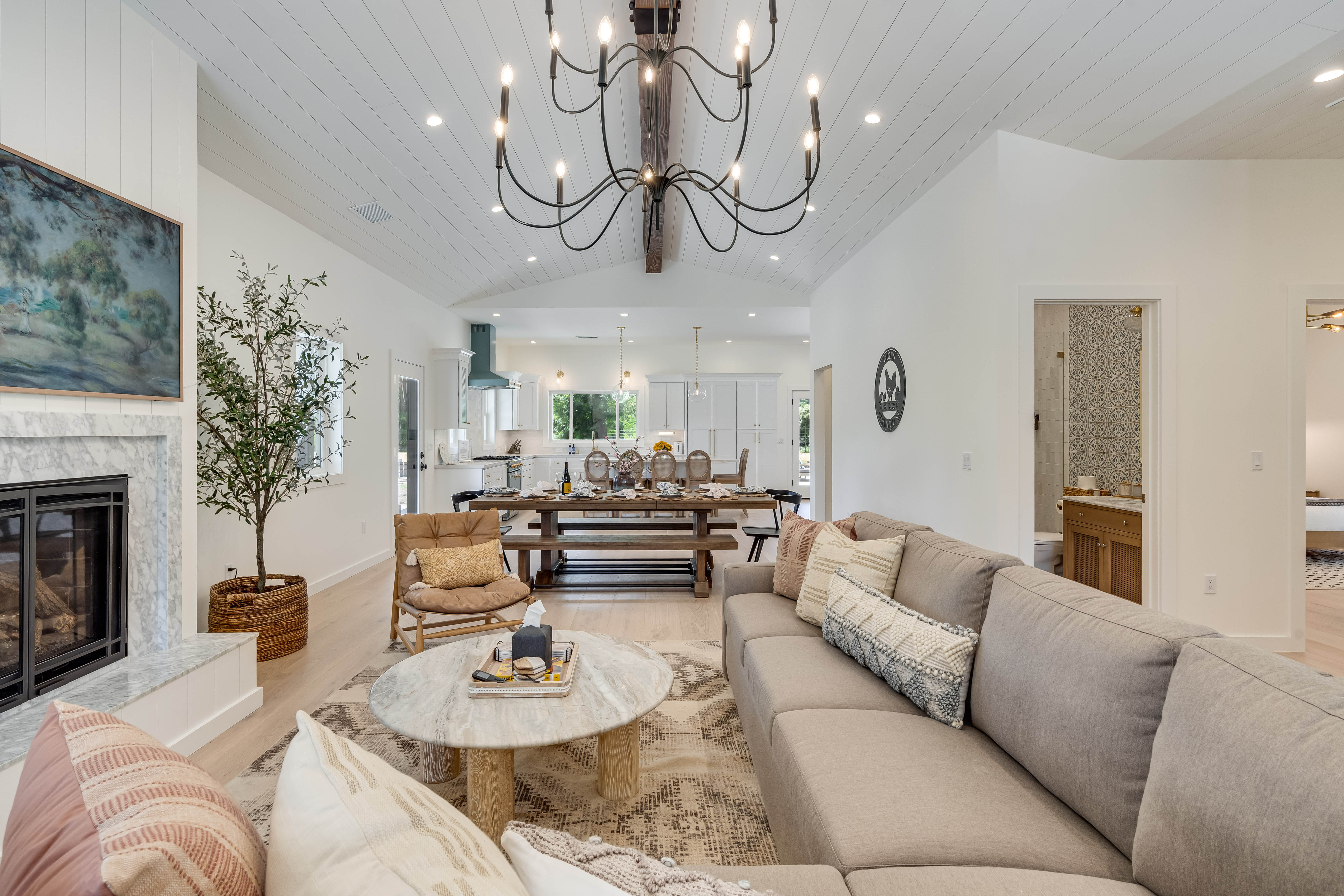 Bright, cozy open-concept modern farmhouse living room and kitchen with vaulted shiplap ceiling, black chandelier, gray sectional sofa, marble fireplace, round coffee table, and long wooden dining table.