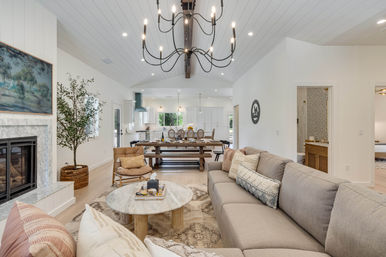 Bright, cozy open-concept modern farmhouse living room and kitchen with vaulted shiplap ceiling, black chandelier, gray sectional sofa, marble fireplace, round coffee table, and long wooden dining table.
