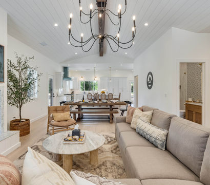 Bright, cozy open-concept modern farmhouse living room and kitchen with vaulted shiplap ceiling, black chandelier, gray sectional sofa, marble fireplace, round coffee table, and long wooden dining table.