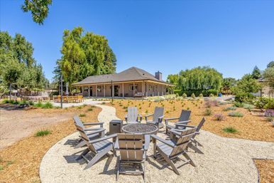 Sunny suburban backyard with circular gravel fire pit surrounded by gray Adirondack chairs, a winding path to a covered ranch-style house, outdoor dining patio, drought-tolerant landscaping and mature trees under a clear blue sky.