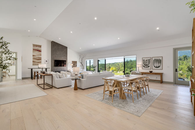 Bright open-concept living and dining room with vaulted ceilings, light oak floors, white sofas around a dark gray fireplace, wooden dining table and chairs on a rug, and sliding glass doors revealing a lush green hillside view.