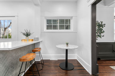 Bright modern kitchen breakfast nook with white marble countertop peninsula, two wooden bar stools, small round marble table with a succulent and dark hardwood floors.