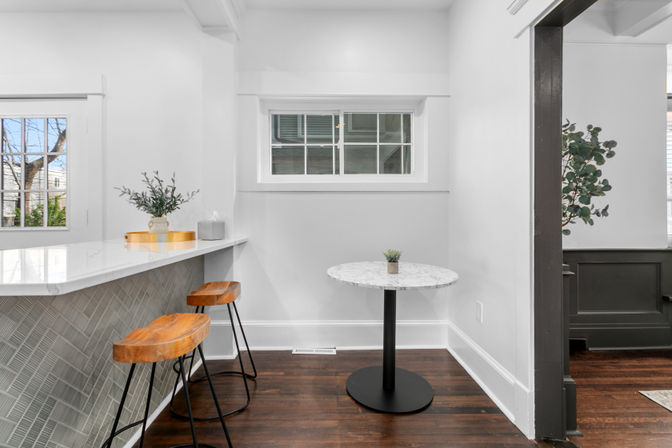 Bright modern kitchen breakfast nook with white marble countertop peninsula, two wooden bar stools, small round marble table with a succulent and dark hardwood floors.