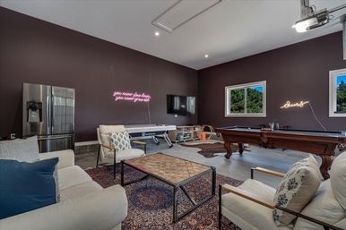 Spacious home game room with wooden pool table, air hockey table, stainless steel fridge, wall-mounted TV, cozy sofas and armchairs on a patterned rug, colorful neon wall signs, and windows showing green trees.