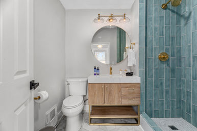 Bright modern bathroom with teal subway-tile shower and brass fixtures, round mirror, wood vanity with marble countertop, white toilet and light marble floor