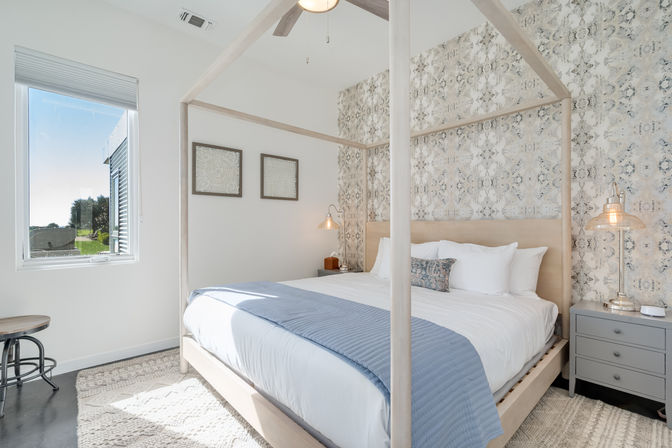 Sunlit modern bedroom with light-wood canopy bed, patterned accent wall, crisp white linens and blue throw, bedside pendant lamps and a window overlooking a green lawn.
