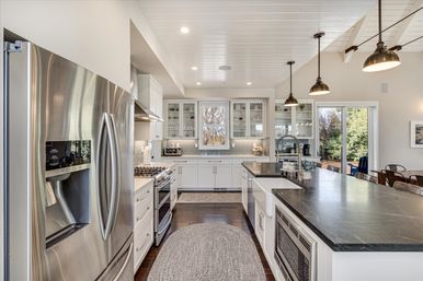 Bright modern farmhouse kitchen — sunlit open-concept space with a long dark stone island, farmhouse sink, stainless steel French-door refrigerator, gas range, white cabinets with glass-front uppers, pendant lights, and sliding glass door to the backyard.